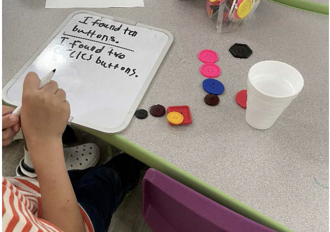 This image shows a child's hand holding a marker. On the table in front of the child is a small white board with writing on it, and several buttons of different colors and sizes. 