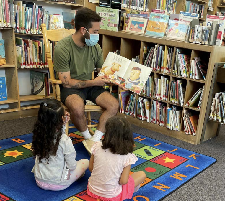 In the Hamden Library, a man wearing a mask reads aloud to two little girls who sit on a colorful rug in front of him.